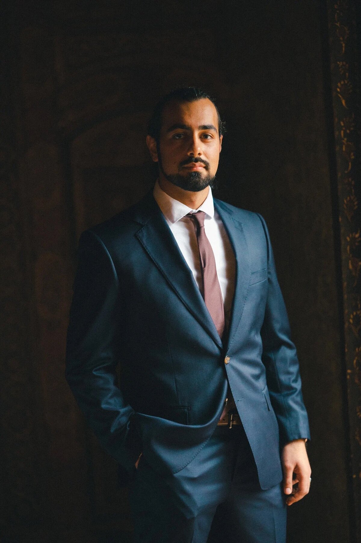 A groom in a blue suit standing confidently, his hands in pockets, against a richly textured dark background, exuding suave sophistication.