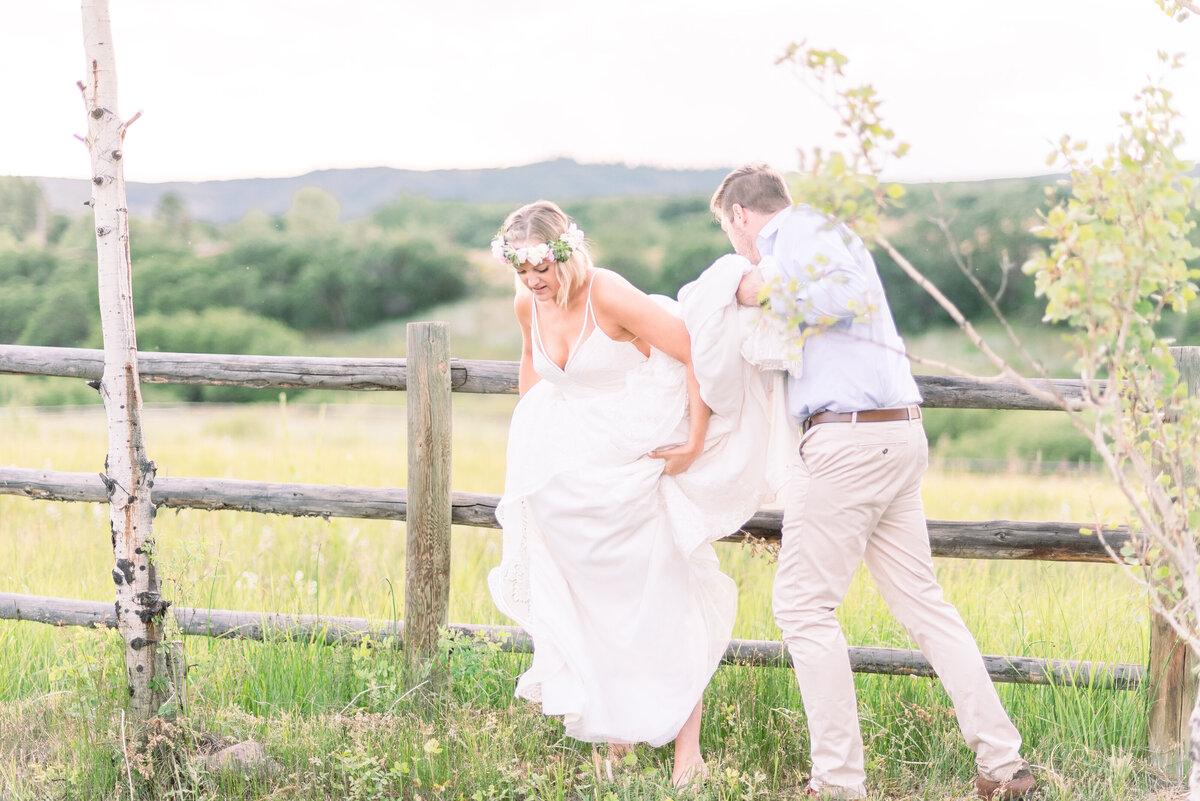 adventurous bride and groom photos with groom helping the bride over a wooden fence in field in Aspen with mountains in the background for a classing Colorado wedding photo in aspen