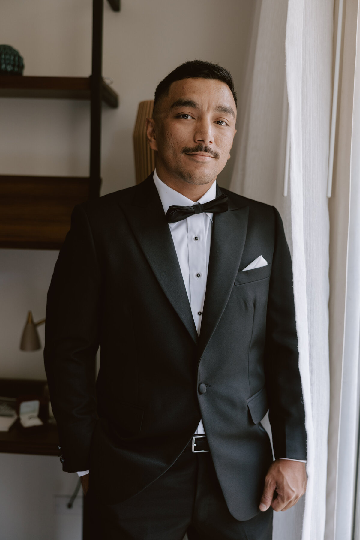 groom portrait looking out a window wearing a tuxedo in laylow waikiki hotel room