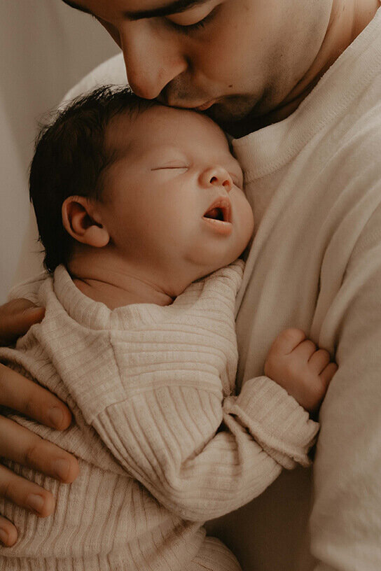 A dad holds his new baby boy against his chest while kissing the top of his head