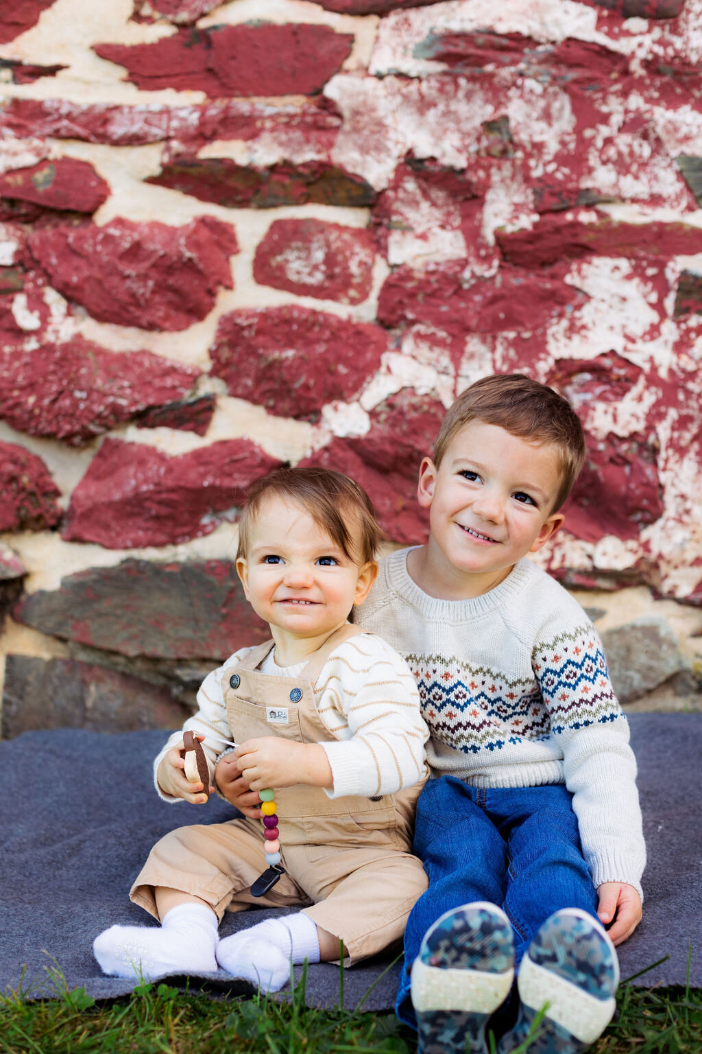 Two children sit outside by a red brick wall.