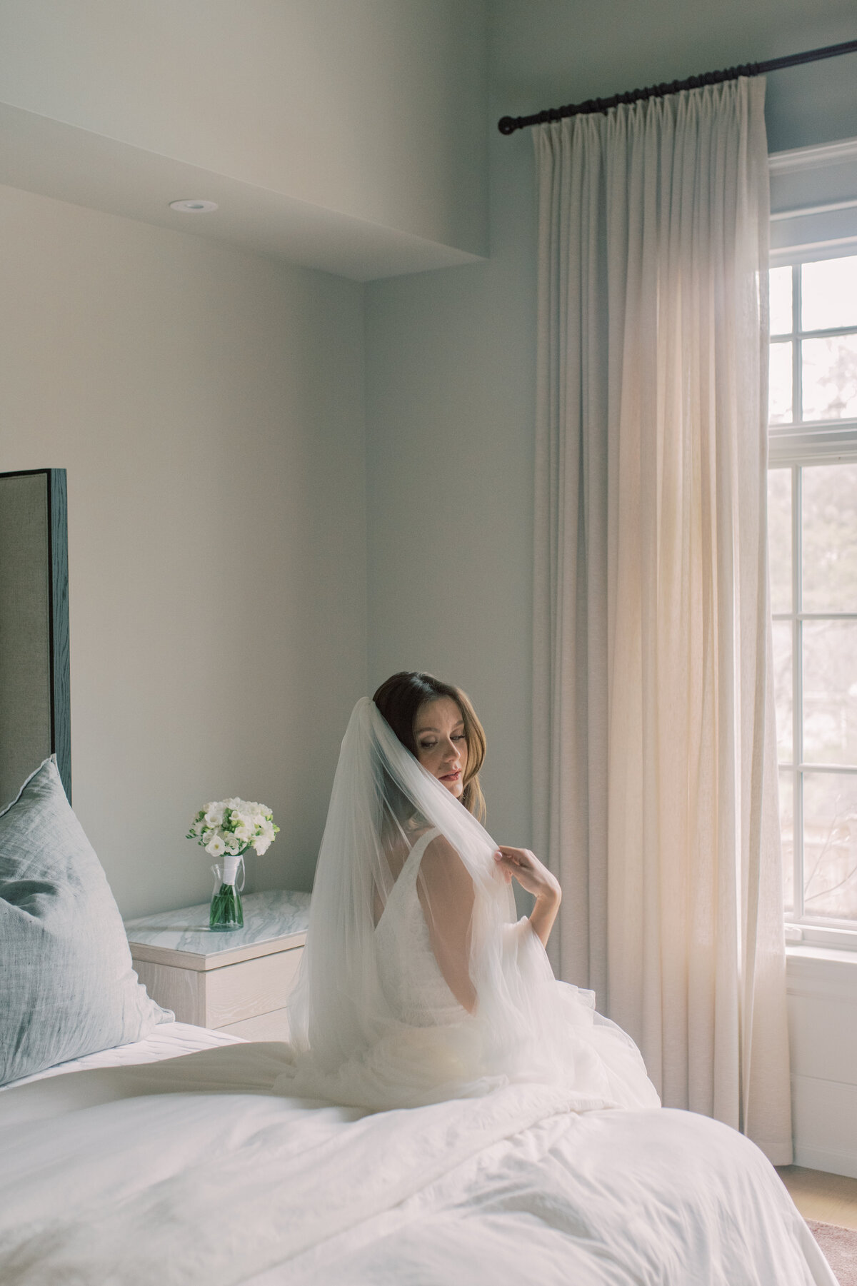 A serene bridal portrait of a pregnant woman sitting on a bed, gracefully holding her veil, bathed in soft natural light through the window