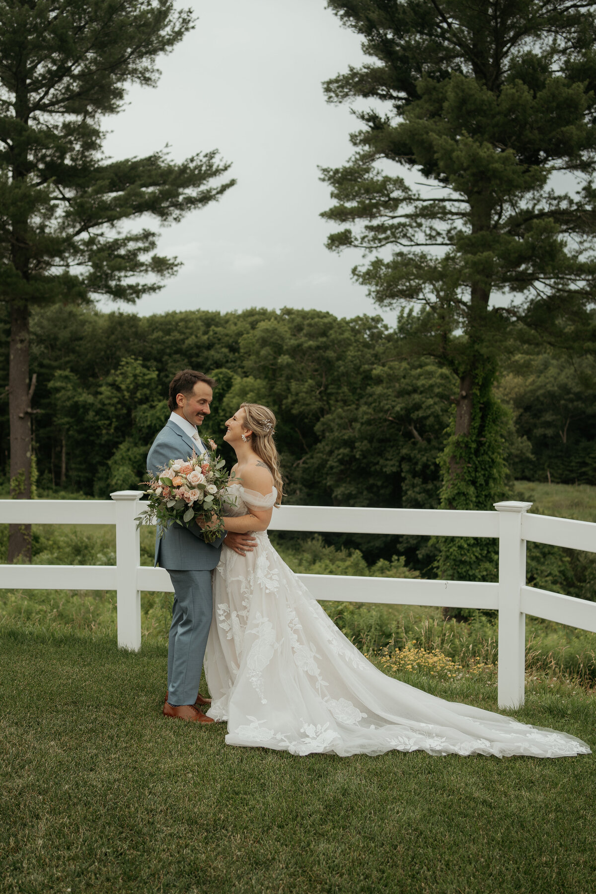 Bride and groom embracing in front of a white fence, surrounded by lush greenery and tall trees. The bride wears an off-the-shoulder lace gown with a long train and holds a bouquet of pastel flowers, while the groom is dressed in a light blue suit. Both are smiling at each other, creating a romantic and sweet wedding moment.