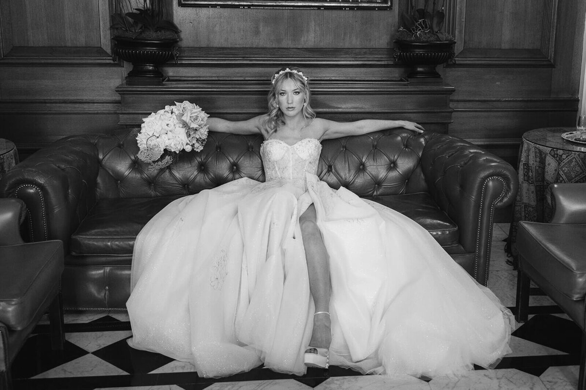 A bride sits carrying a bouquet in her right hand with one leg showing through her dress. She wears Sophia Webster wedding shoes.