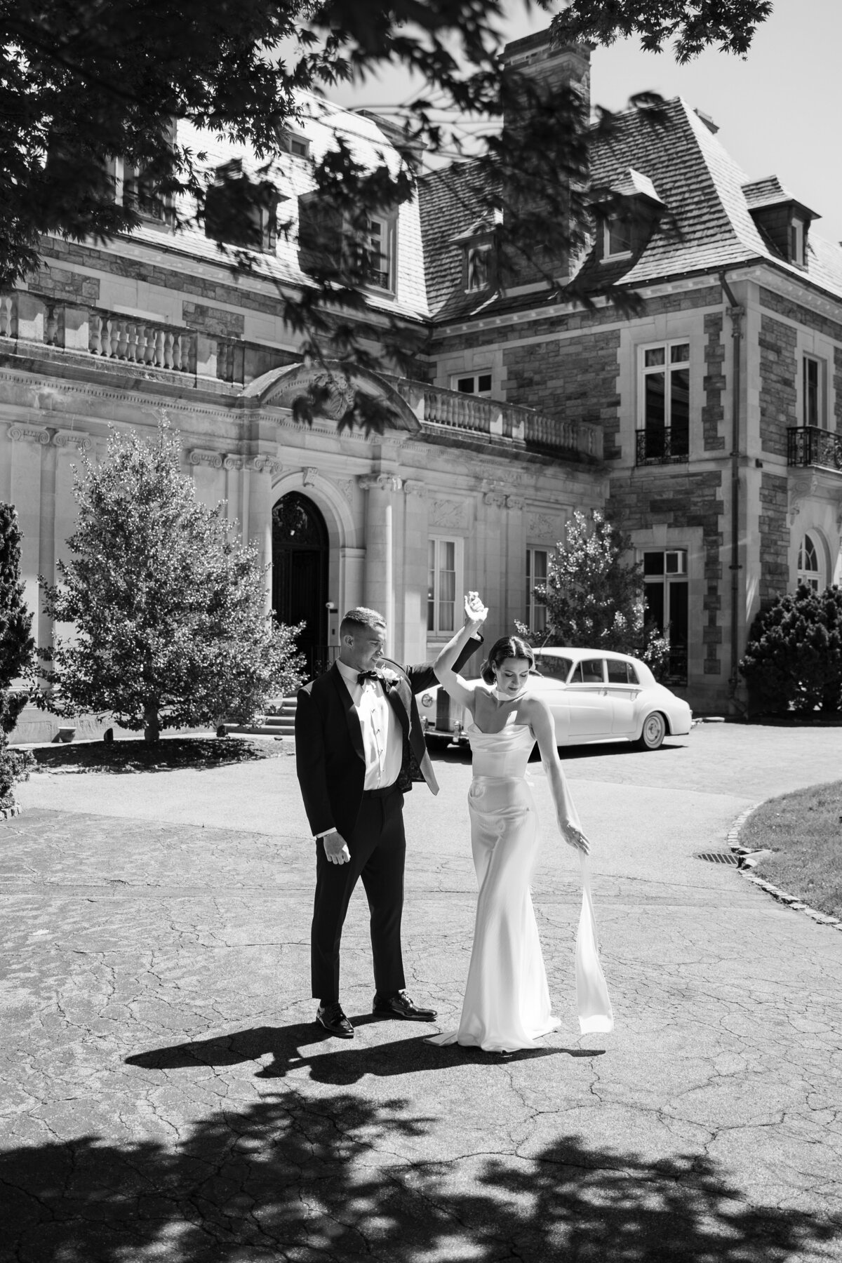 Aldrich Mansion Wedding Photos | Kelsey Sheehan Photography Timeless Rhode Island Weddings | A joyful bride in a flowing gown and groom in a tuxedo pose outside a grand mansion with a vintage car behind them in this black-and-white photo.