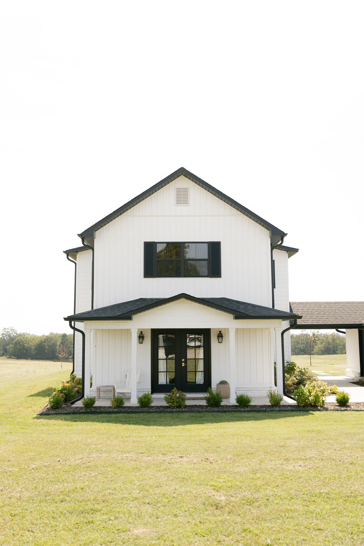 A bright exterior photograph of a modern white farmhouse wedding venue featuring black trim, double black front doors, manicured landscaping, and open fields in the background. Perfect for rustic, countryside, and modern farmhouse weddings.
