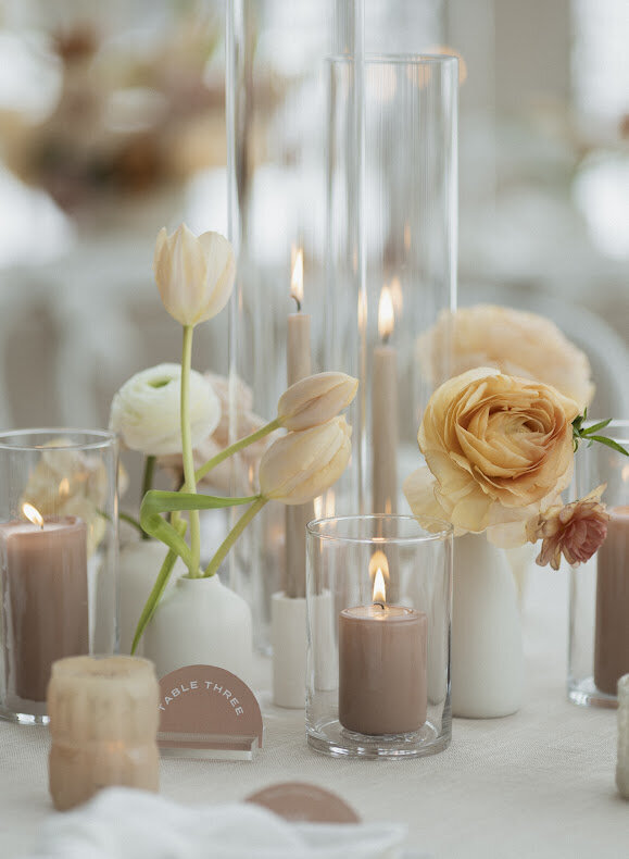 A close-up of a bohemian guest table centrepiece at a National Arts Centre wedding, featuring taupe tapered and pillar candles surrounded by white bud vases with peach, cinnamon and white fresh flower booms and a modern half-circle table number.