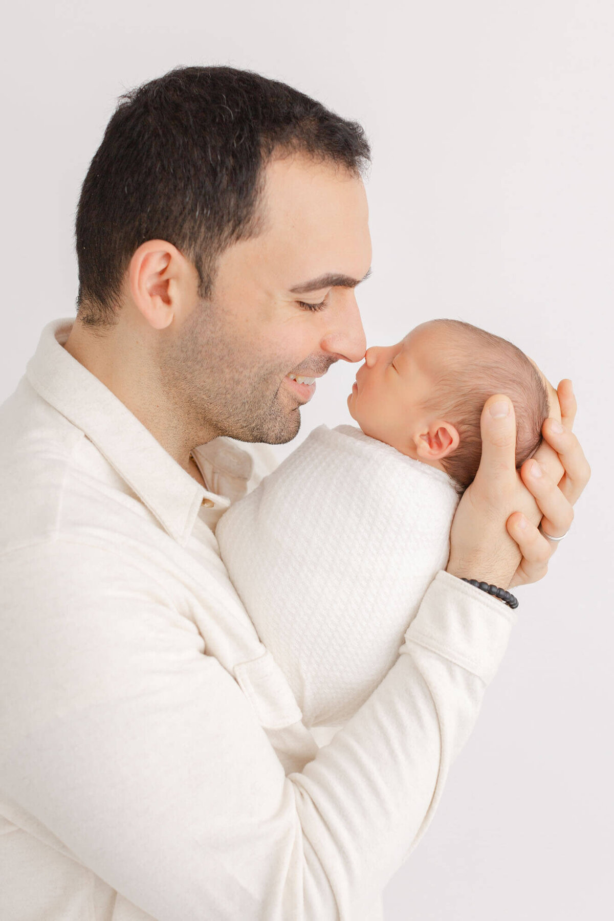 Dad in cream shirt holding baby in arms in front of him and their noses are touching. Dad is smiling and looks so happy. Newborn photos in Portland, Oregon.