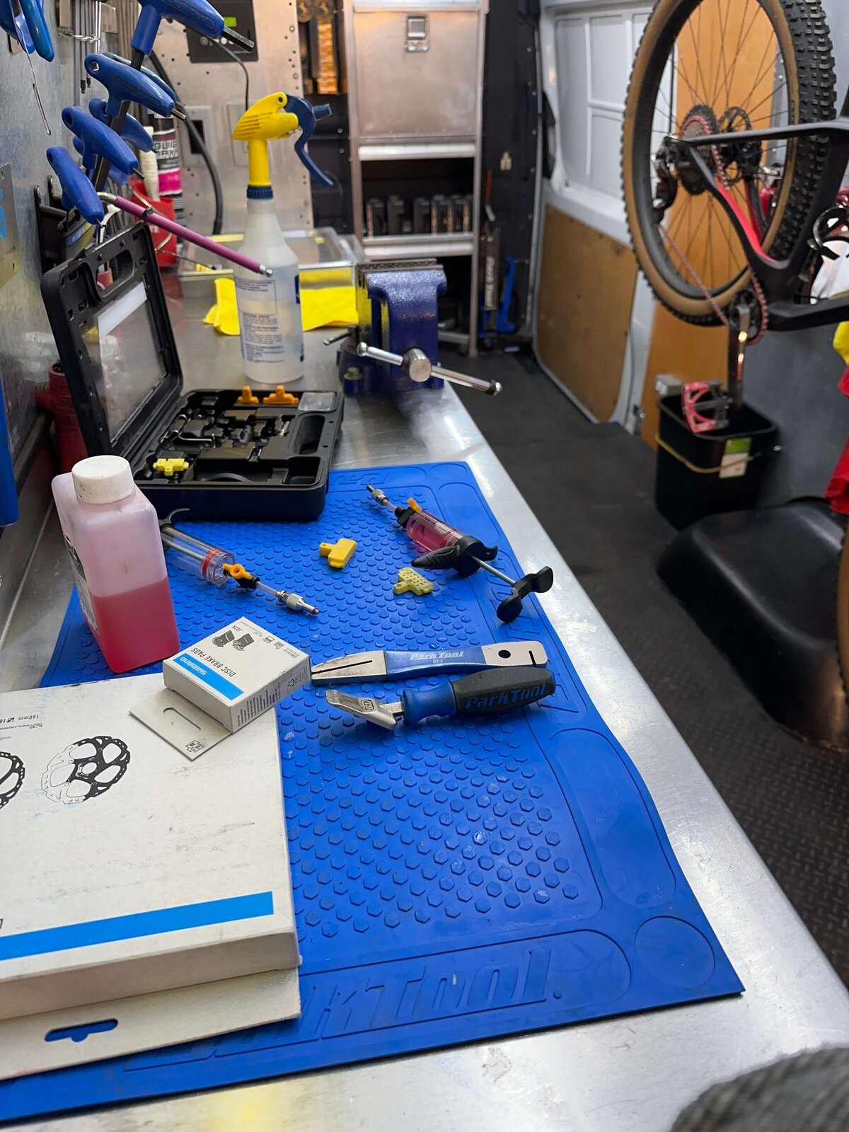A bike repair workspace featuring tools, a toolbox, a spray bottle, and bike parts on a blue mat. A bike is mounted in the background. The setting is inside a workshop with a focus on maintenance and repair