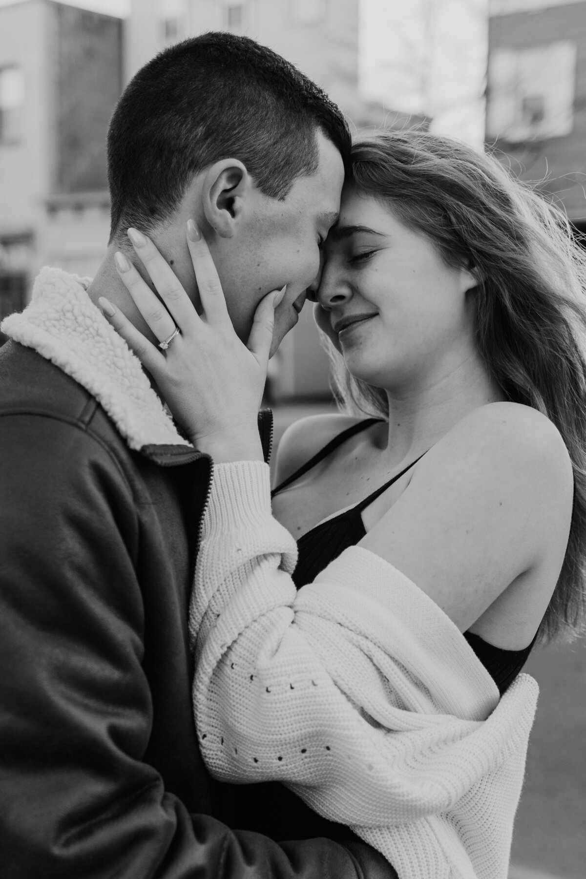 A black and white photo capturing an intimate moment between an engaged couple, with the woman embracing the man's face gently as they touch foreheads, showcasing their affection and the woman's engagement ring.