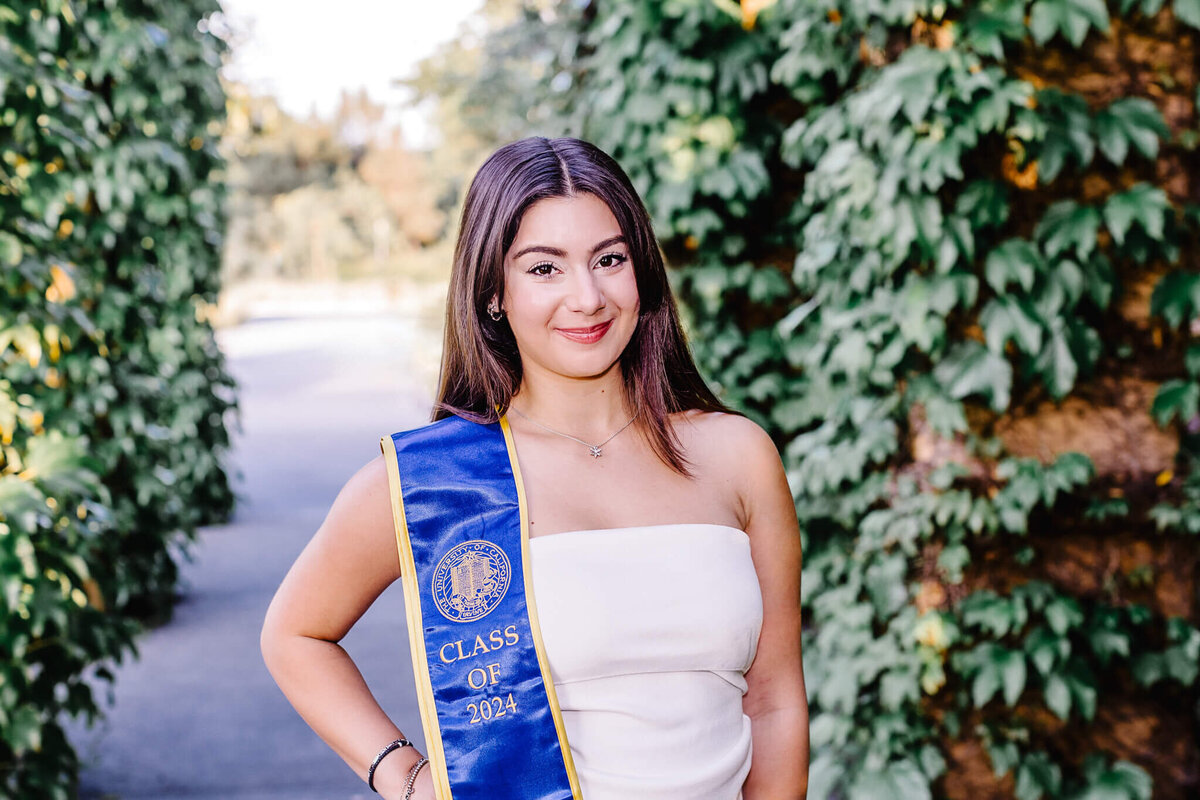 A young woman in a white dress stands outdoors, smiling, with a blue graduation sash that reads Class of 2024. She is surrounded by lush green foliage.