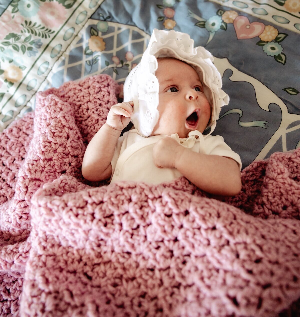 A newborn baby girl wearing a white onesie and bonnet tucked in under a pink knitted blanket in her Edmonton nursery