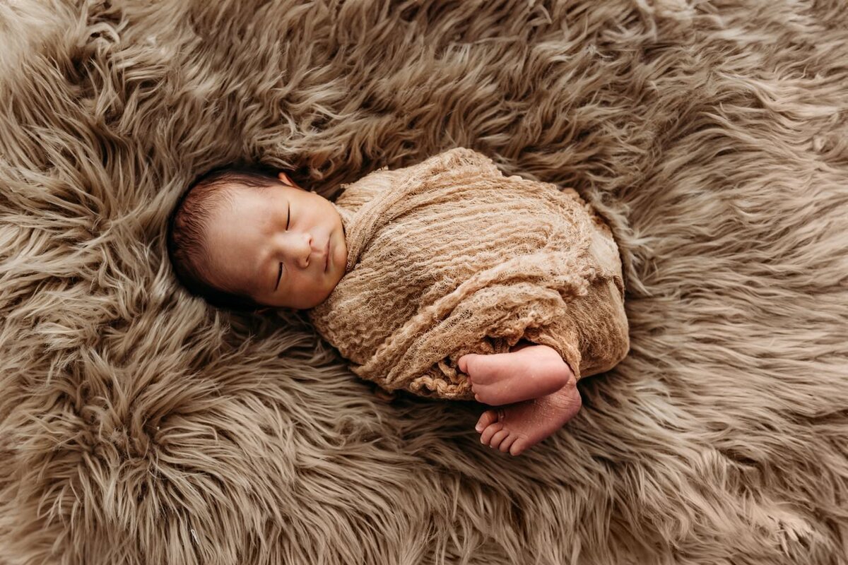 newborn sleeping on a tan fur rug wrapped in a similar colored swaddle. babys feet are sticking out.