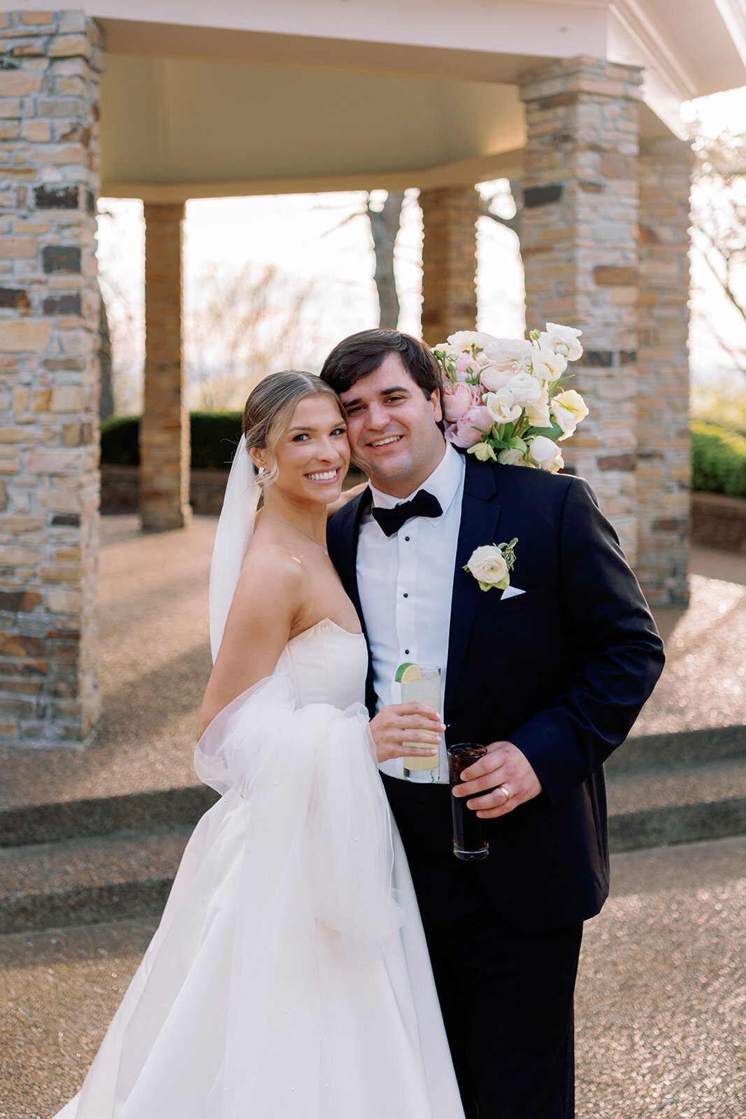 Bride and groom smiling and holding drinks while embracing in front of the stone pavilion at Burritt on the Mountain in Huntsville, Alabama, photographed by Perry Hancock.