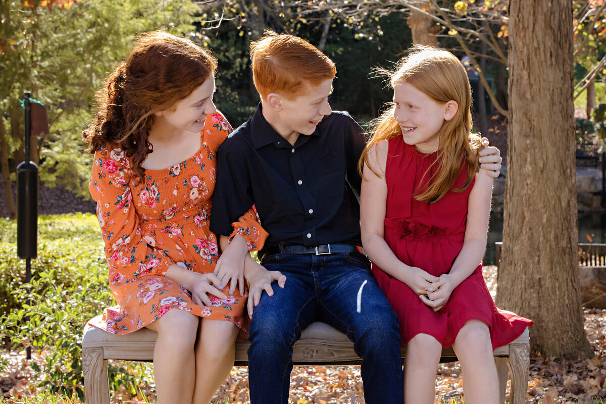 Gleeful sibling portrait of brother and two sisters seated on bench in autumn park at Ballantyne Corporate Park