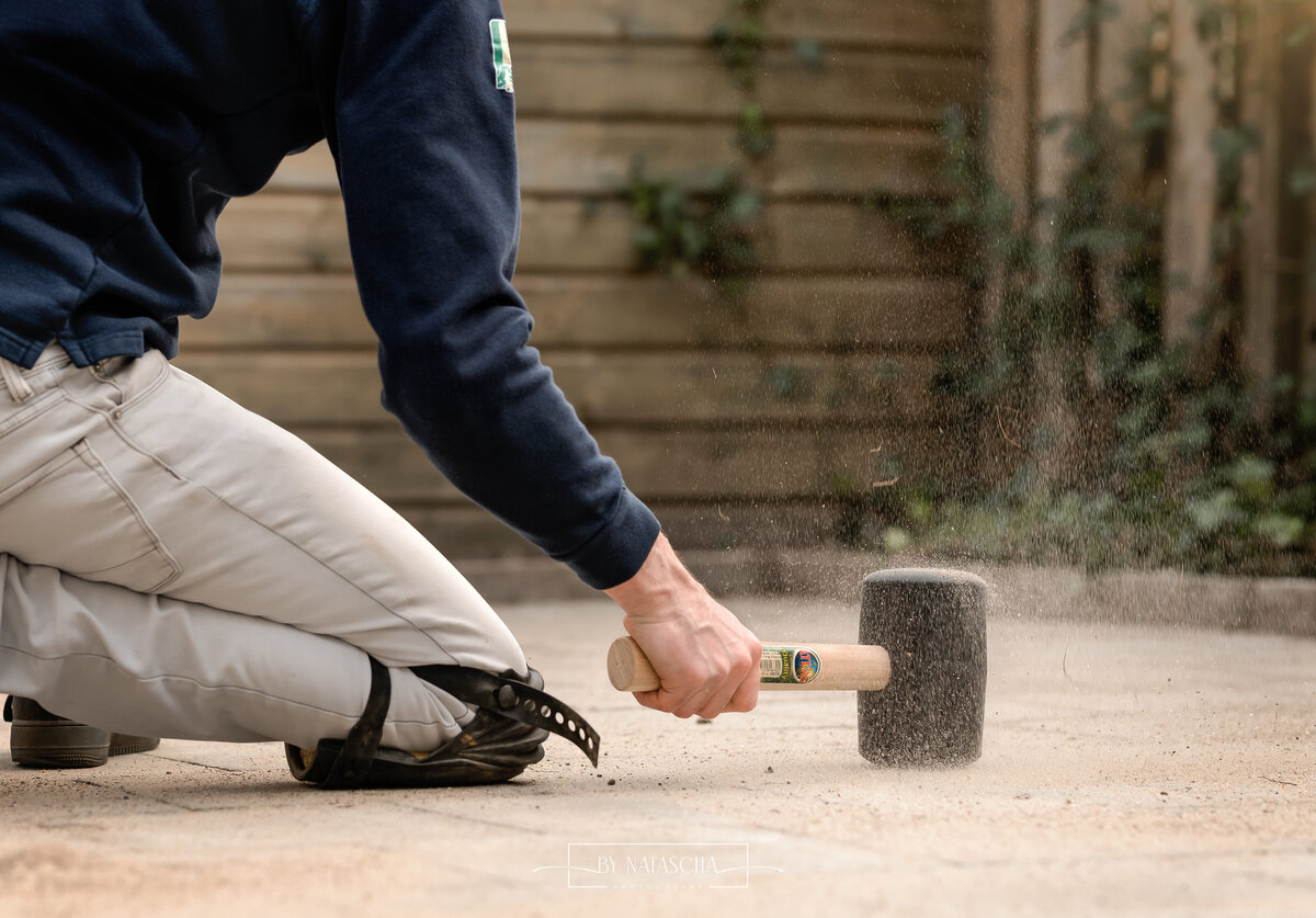 Stratenmaker slaat de tegels aan in de tuin die net gelegd zijn, waarbij hij kniebschermers draagt en een hamer in de hand die tijdens het slaan zorgt voor opspattend zand