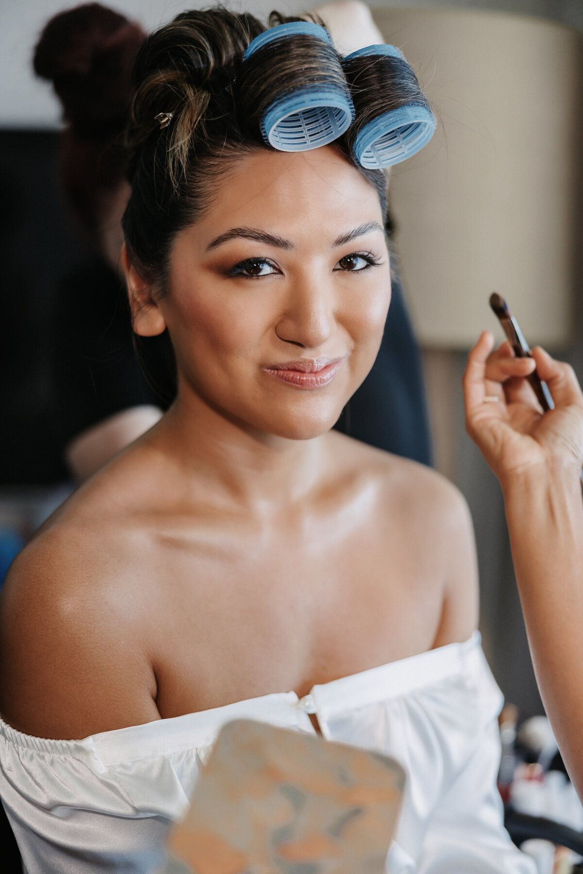 bride with hair in rollers and a hand reaching towards her face with a makeup brush