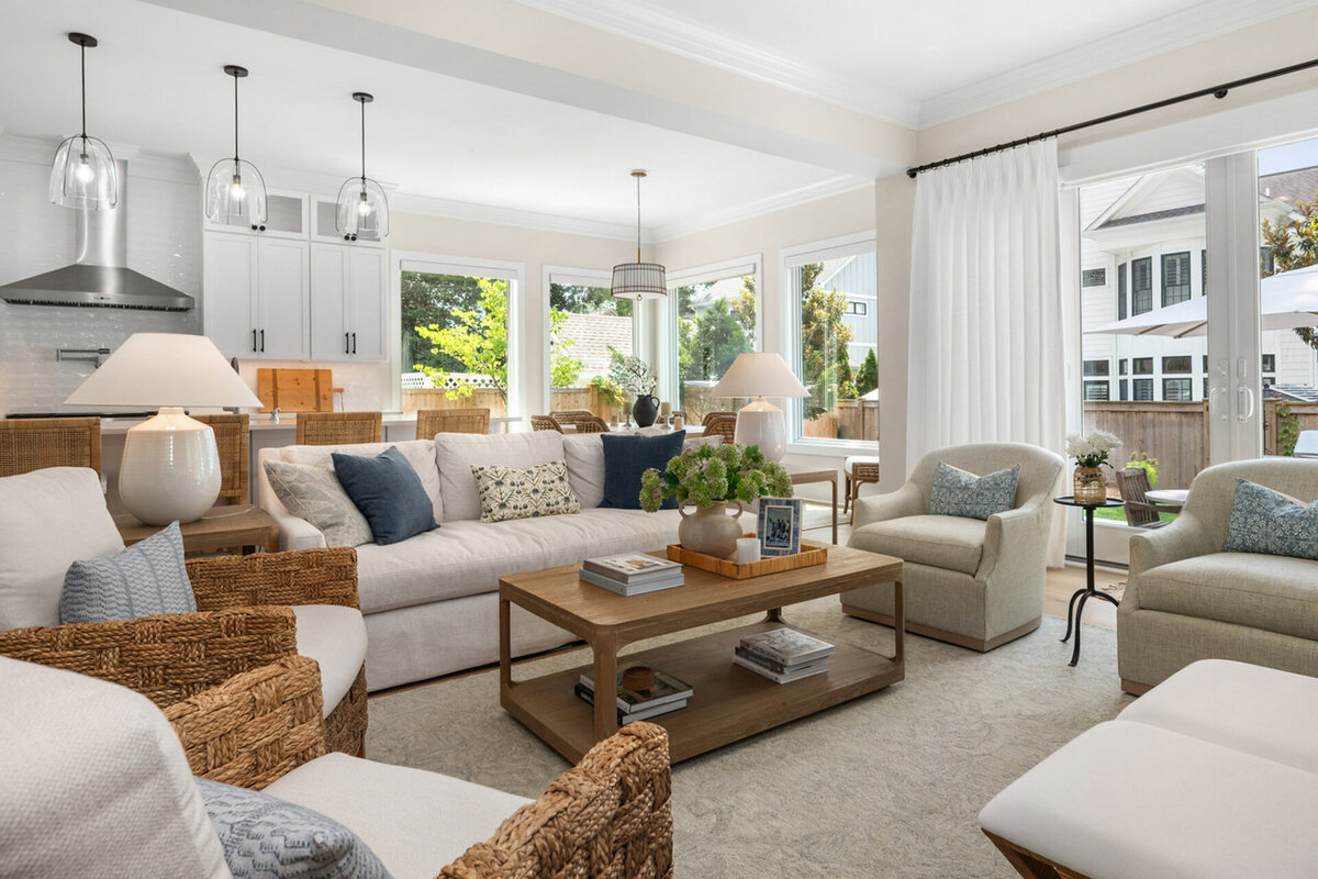 Luxury real estate listing photo featuring a bright living room with white sofas, wicker chairs, and a wooden coffee table. Large windows provide natural light, and a kitchen with pendant lights is visible in the background.. Photographed by Real Estate Photographer Svetlana Leahy of Refined Capture   , based in Ocean City, MD