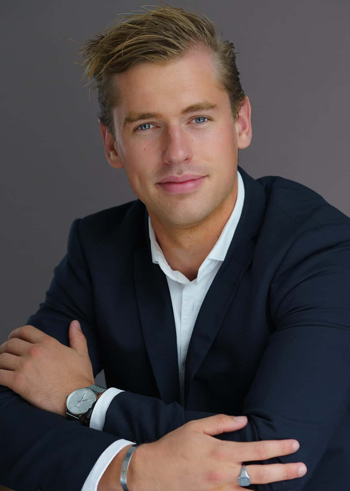 Man in a dark suit with a white dress shirt sporting a silver watch sitting for a photo