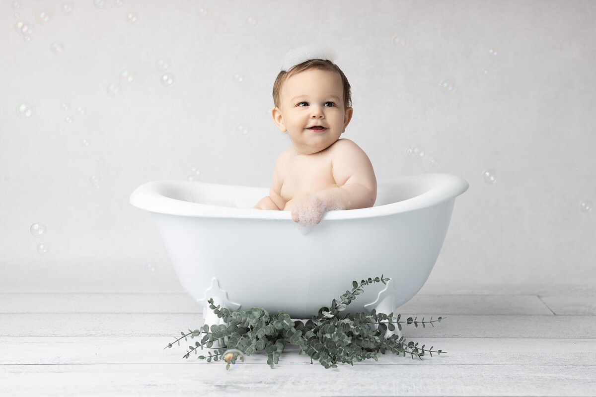 Dallas milestone photographer captures a sweet baby in a white vintage tub, bubbles in the air and suds resting playfully on the baby’s head.