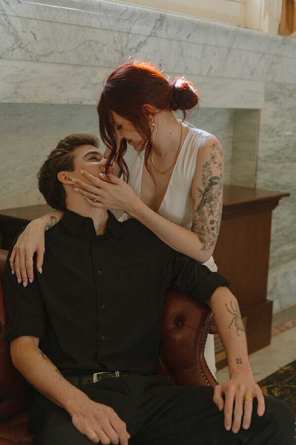 A woman looks down and holds the face of her husband, whose sitting down on a red leather couch.
