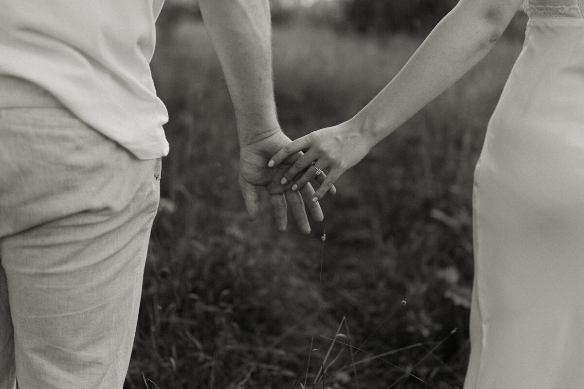 An engaged couple brushes finger tips while walking through tall summer grass.