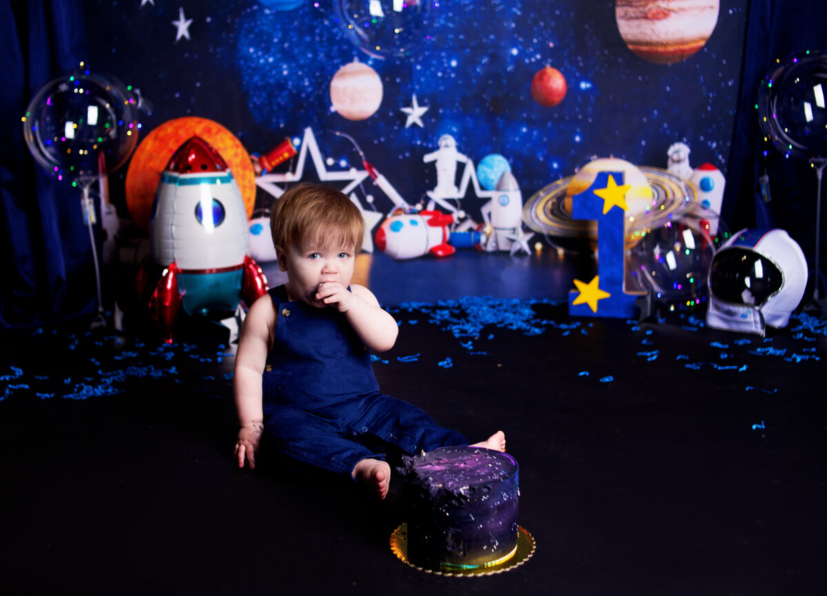 Baby boy wearing blue overalls eating cake during irst birthday cake smash photo shoot in Franklin Tennesse photography studio, with Franklin TN Family photographer
