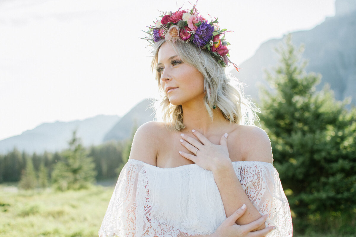 Stunning bride wearing an off-the-shoulder lace wedding gown, large floral crown of purple and pink, in the mountains, captured by Christy D. Swanberg Photography, editorial elopement and wedding photographer in Calgary, Alberta, featured on the Bronte Bride Vendor Guide.