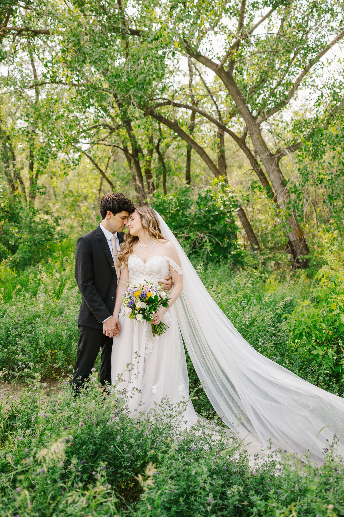 Bride and groom standing with their heads together in front of a wooded area