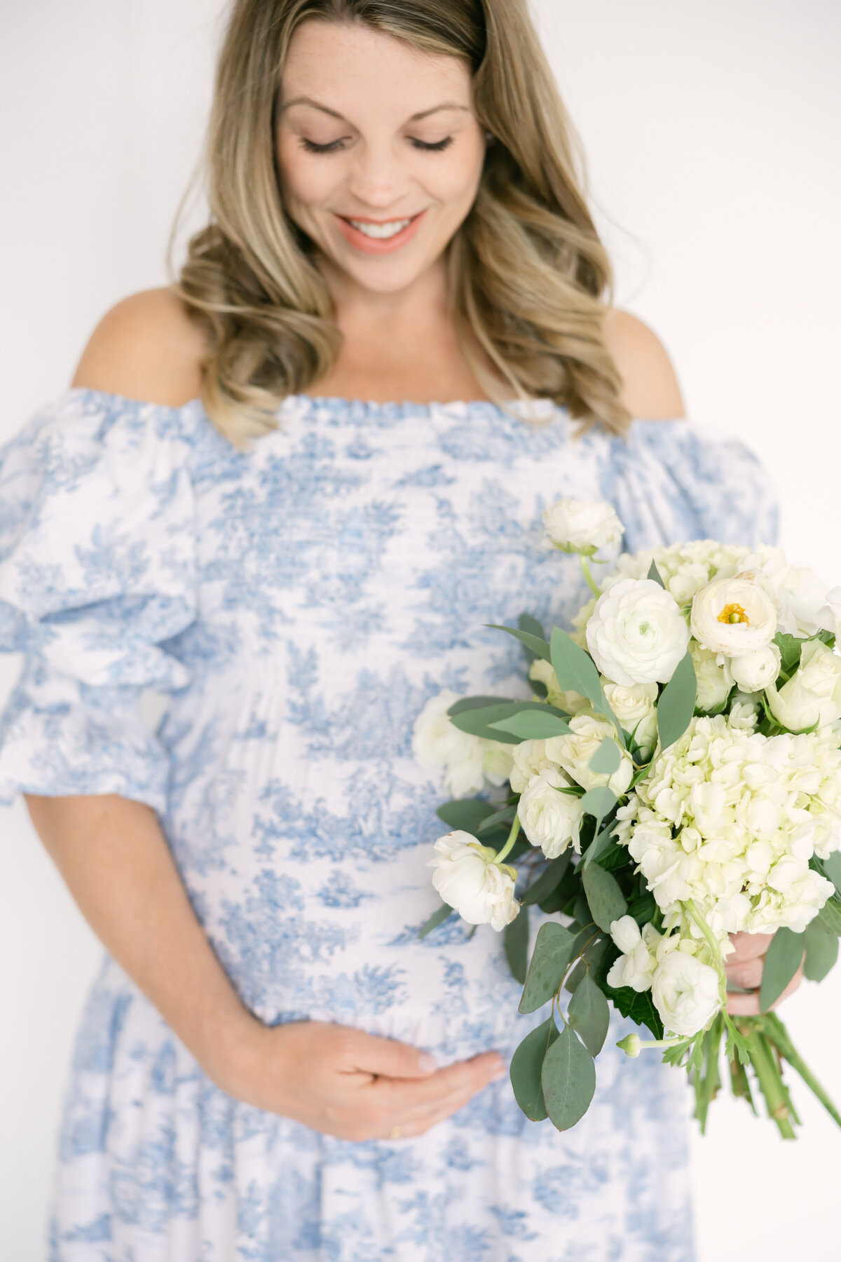 a mother smiles down at her pregnant belly as she wears a blue maternity dress and holds a white floral bouquet