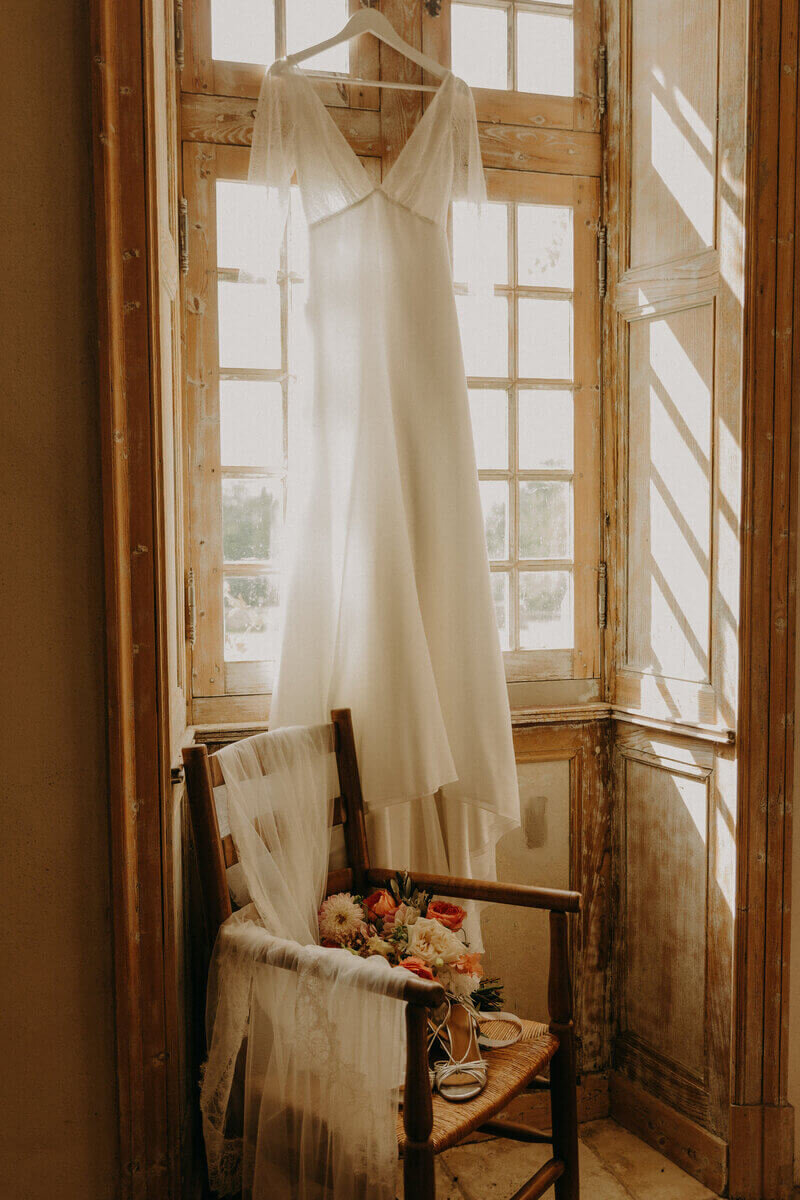 Robe blanche suspendue à un cintre accrochée à une fenêtre en bois. Dans l'encadrure une chaise en bois avec le bouquet et les chaussures de la mariée. Photo prise pendant la séance photo mariage en Vendée.