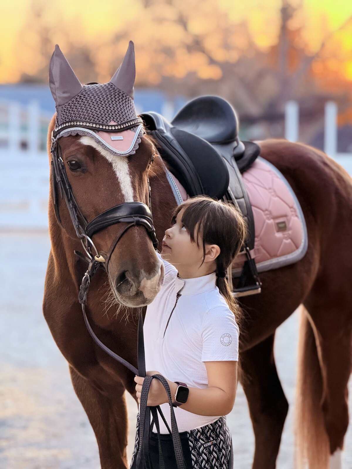 A young girl looking sweetly at a chestnut pony, the pony has a pink saddle pad and ear bonnet