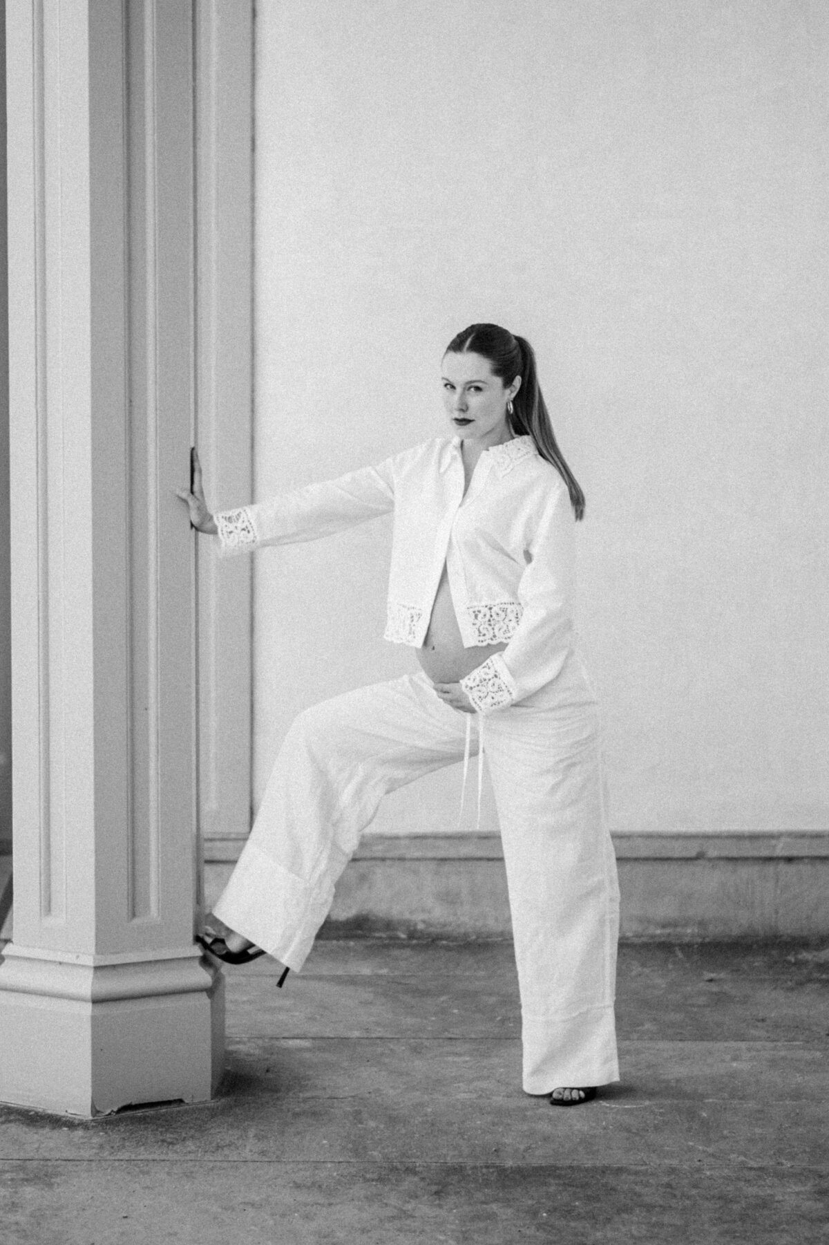Editorial black-and-white maternity portrait of a poised mother-to-be in heels, framed by architectural columns