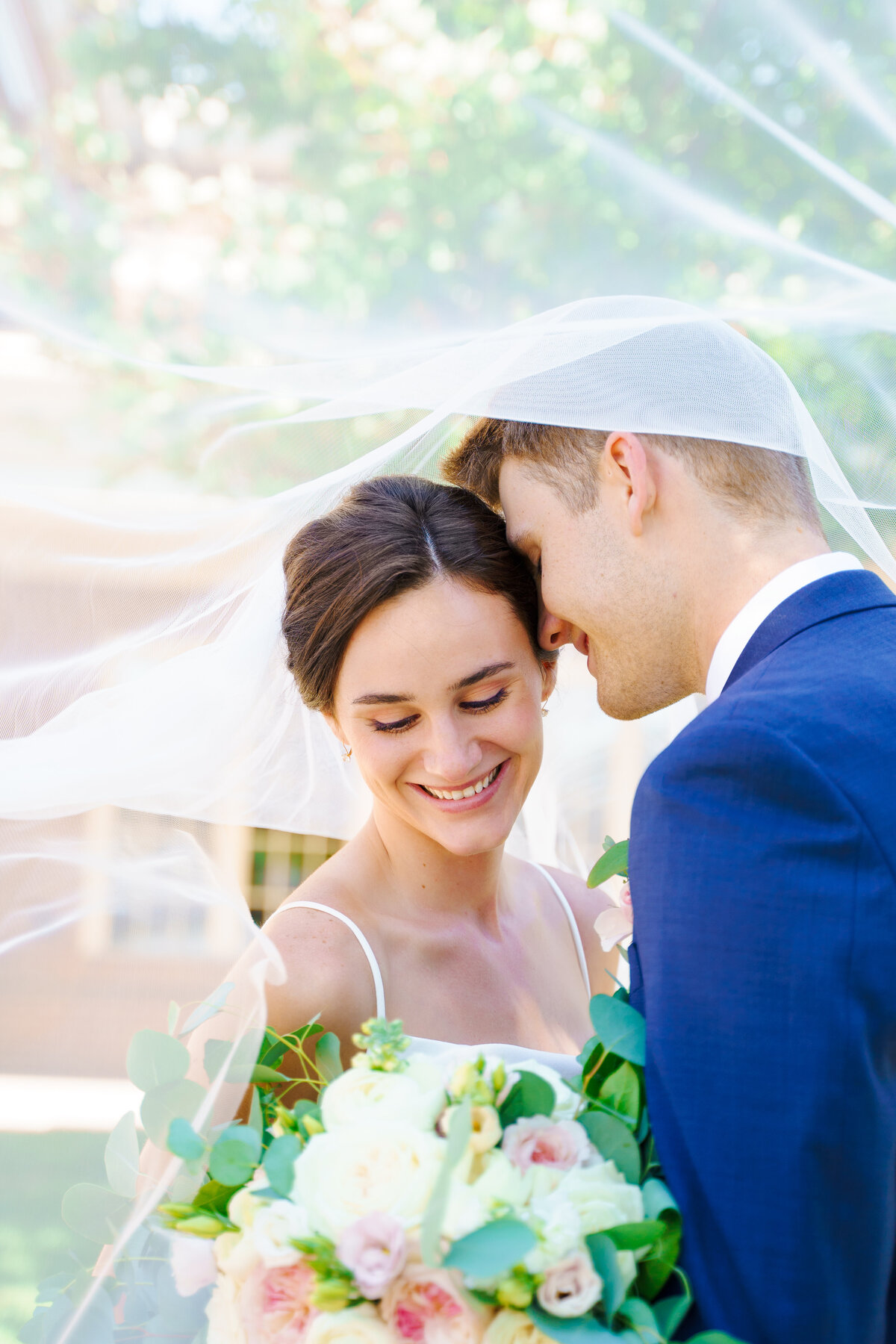 Bride smiles down at her bouquet as her groom nuzzles into her temple while they're underneath her veil at their wedding at The Estate at New Albany.