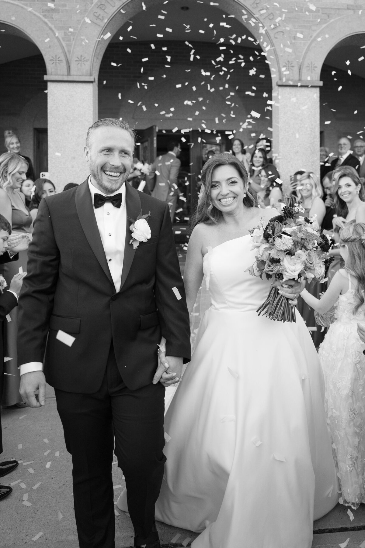 A bride and groom smile and laugh as they walk through confetti. 