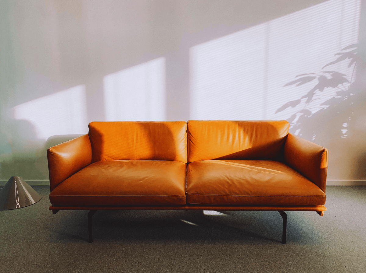 Egy elegáns, barna bőr kanapé áll egy világos szobában. A kanapé előtt egy modern lámpa látható, a falon pedig érdekes fény- és árnyékjáték figyelhető meg.