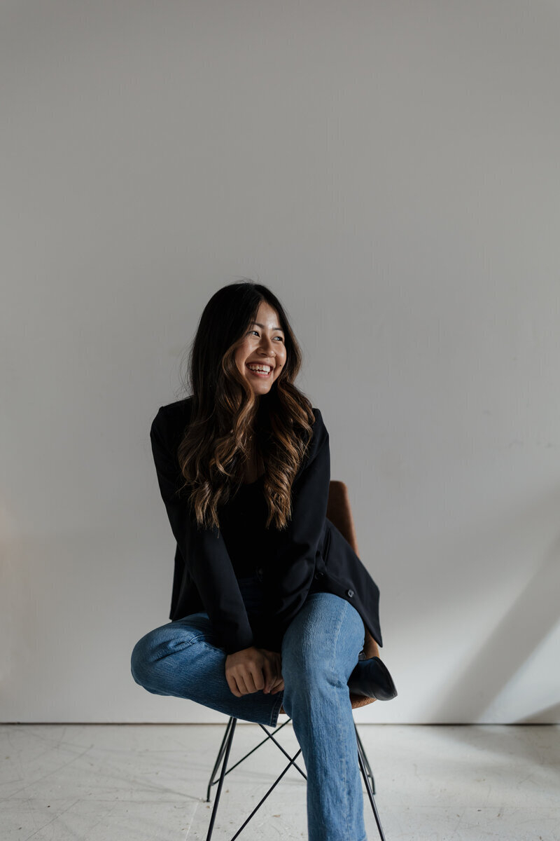 A person with long dark hair, wearing a black top and blue jeans, sits on a chair against a plain white background. Captured by a New England Portrait Photographer, they smile while gazing slightly to the side, exuding effortless charm in every frame.