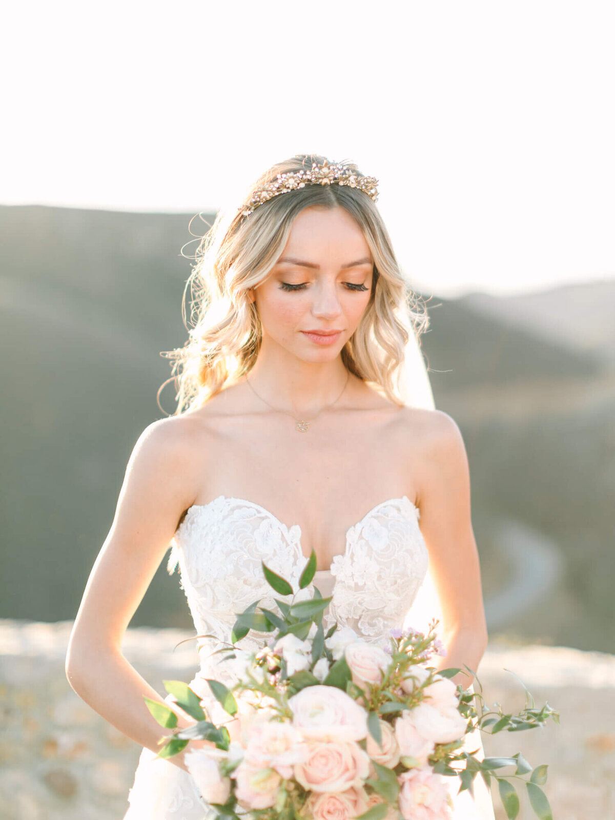 A bride in a strapless lace gown holds a bouquet of pink roses and greenery, with soft sunlight and blurred hills in the background.