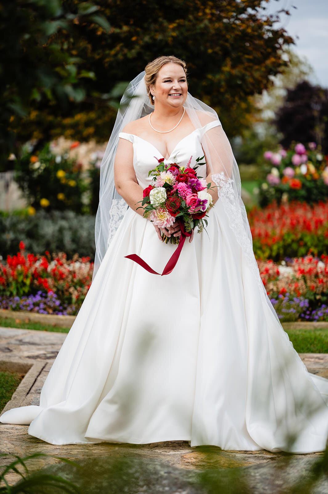 A bride in a white gown with a long train and veil holding a bouquet, smiling widely in a garden setting