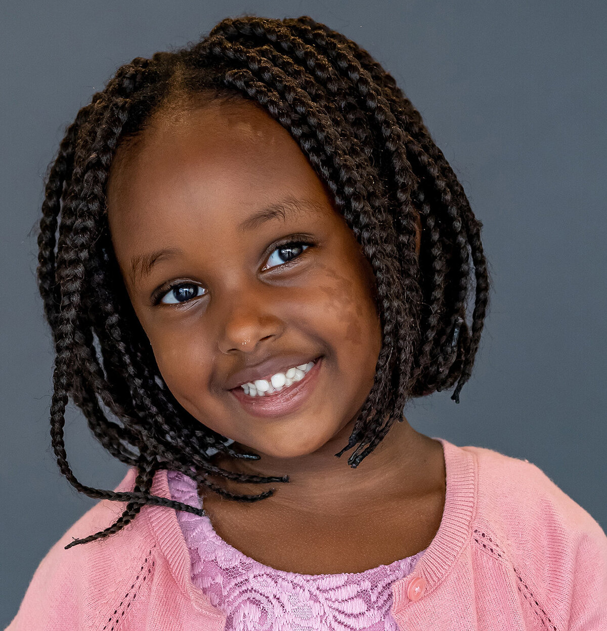 Preschool girl, hair in braids and wearing a pink dress smiles  for her school photos in port coquitlam, BC.
