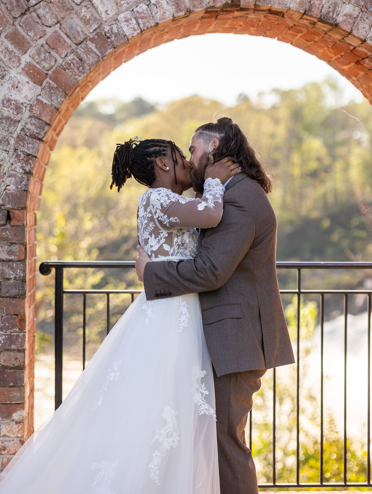 black bride wearing a lace dress and white groom wearing brown suit  kissing with waterfall behind them at The Bibb Mill by Columbus Georgia wedding photographer Amanda Richardson Photography