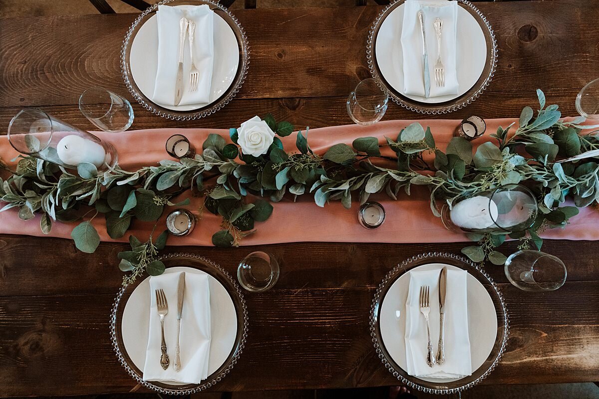 A dark barn wood farm table set with clear glass beaded chargers and white plates, napkins and flatware placed in the center. The table has clear glass cylinders with white pillar candles, a terracotta table runner and a garland of eucalyptus greenery.
