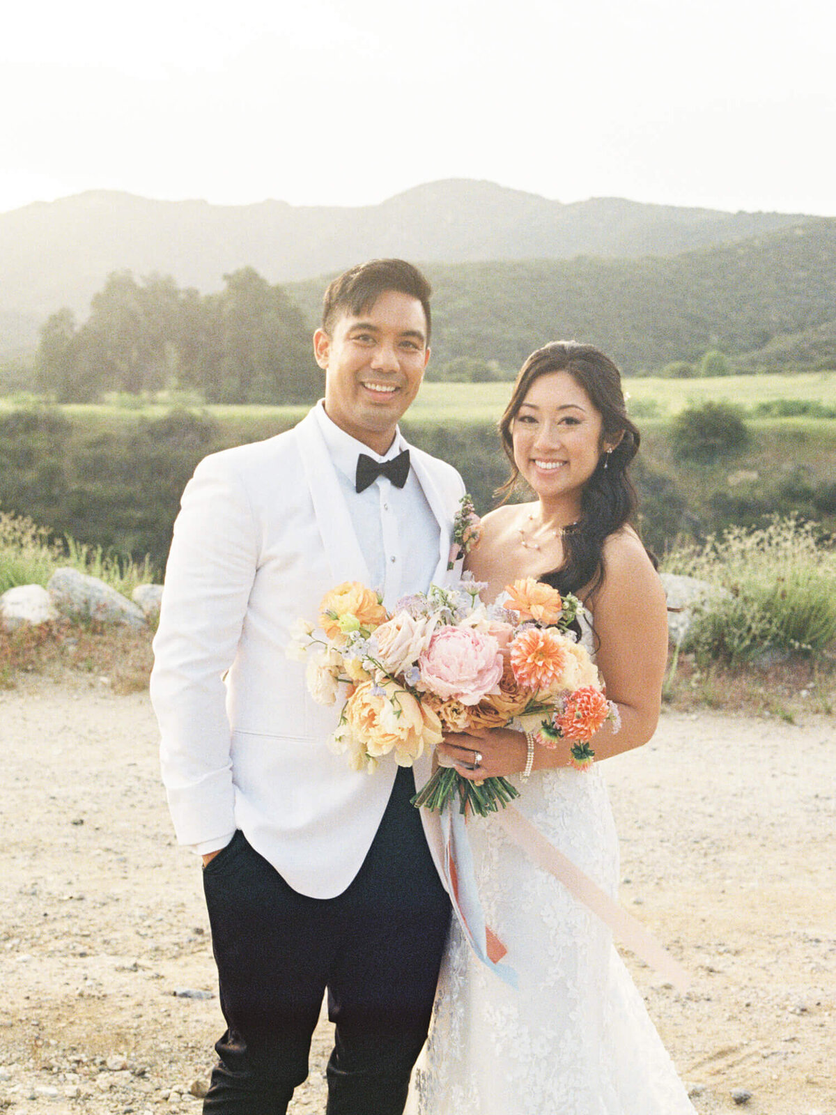 A smiling couple in wedding attire stands outdoors against a backdrop of mountains. The bride holds a bouquet of colorful flowers. The scene is bright and joyful.