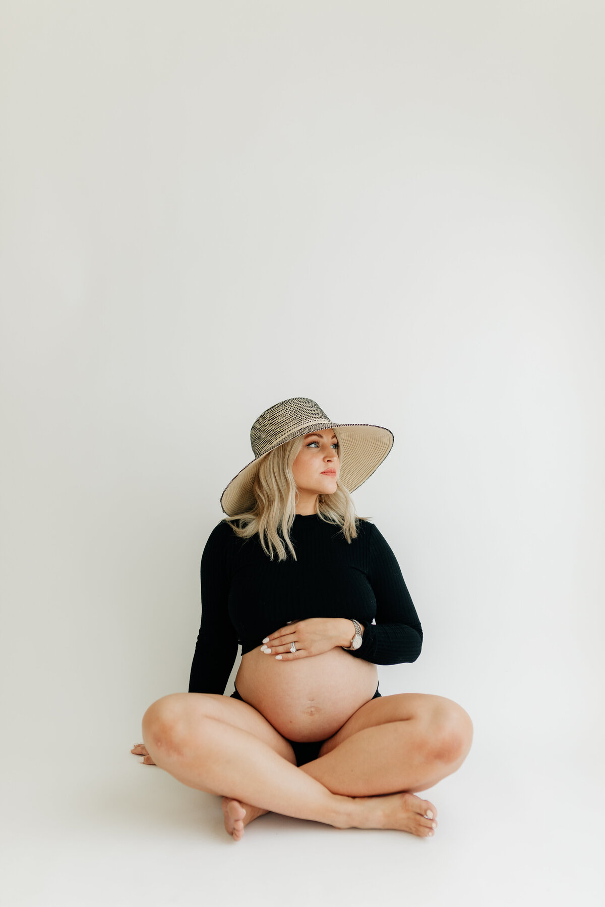 Pregnant woman sitting on ground in white studio setting, showcasing her bare pregnant belly with a large hat on 