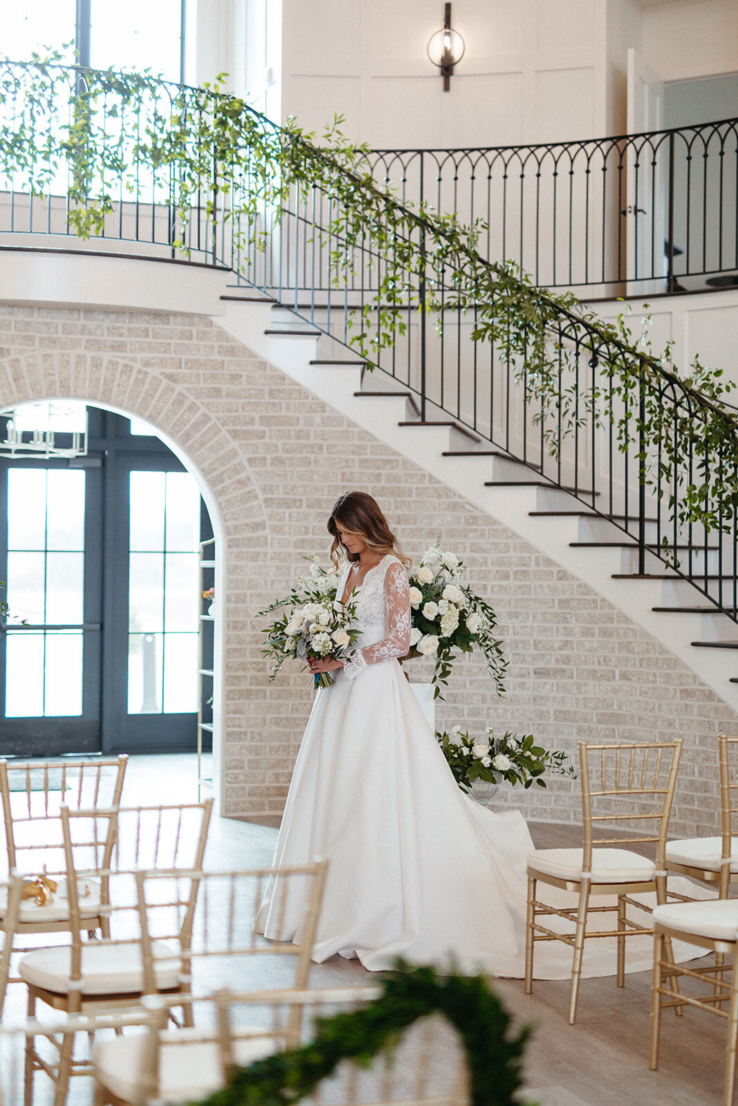 Bride in a white wedding gown holds a bouquet while waiting at the altar in front a staircase wrapped in garland.