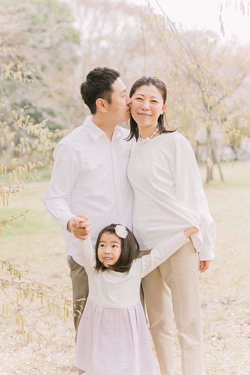 パパがママの頬にキスしていて、娘の女の子が二人の前でおどけている可愛らしい家族写真。相模原市の公園で撮影。