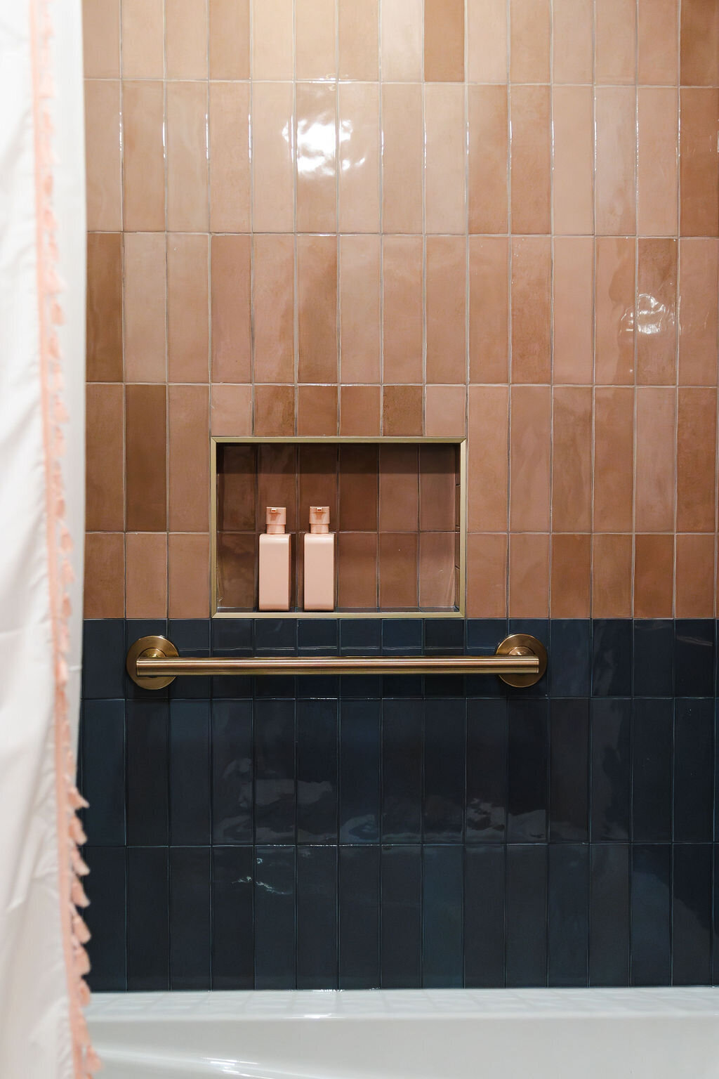 A detail of a bathtub and shower area with a combination of dark blue and warm brown vertical tiles. The lower section of the wall is tiled in dark blue, while the upper section features a mix of light and medium warm brown vertical tiles. A horizontal brushed gold grab bar is mounted on the dark blue tiles. A rectangular niche within the warm brown tiled area holds two small, light pink bottles. The edge of a white bathtub is visible at the bottom, and the edge of a white shower curtain with a delicate pink trim is on the left. The tiles have a slightly glossy finish, reflecting the light.