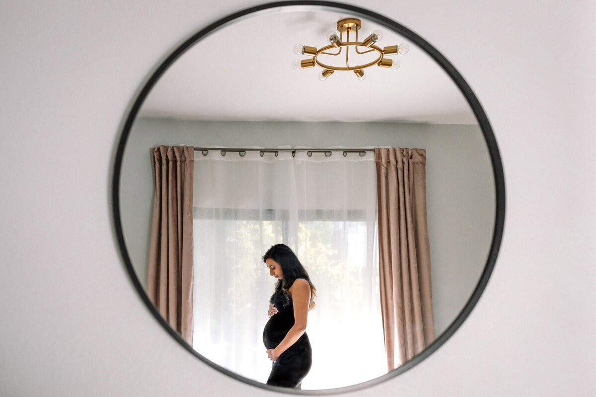 reflection in mirror of pregnant woman in black dress holding her baby bump and standing in front of window with curtains