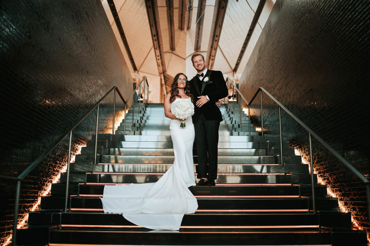 A couple elegantly poses on illuminated stairs at a modern venue, showcasing a timeless wedding moment.