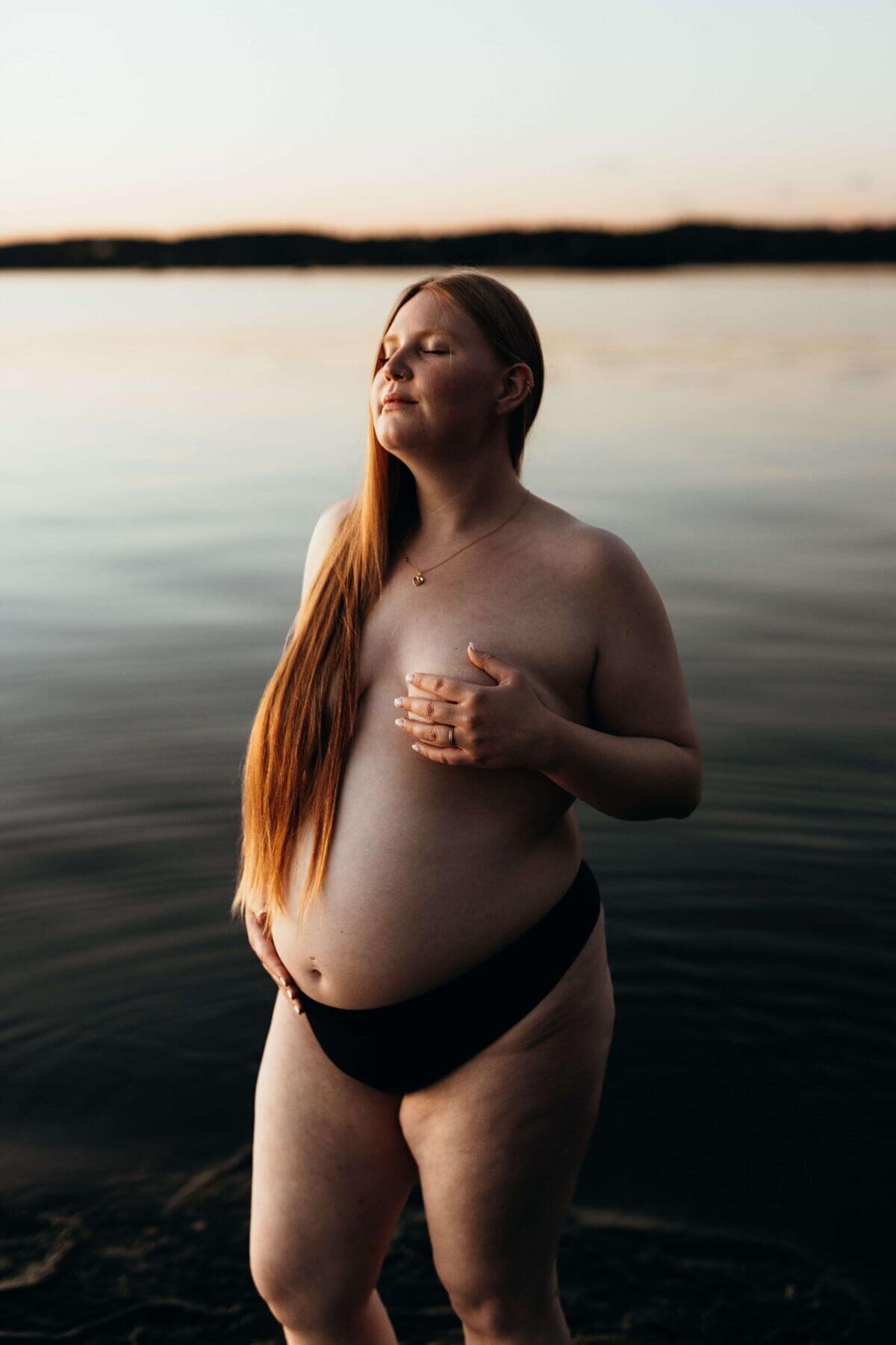 En gravidbild på en gravid kvinna med långt orangeat hår ståendes vid vattnet iklädd svarta trosor under en gravidfotografering i Stockhol